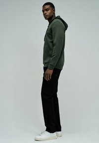 Sudadera verde oscuro con un bolsillo frontal y cordón, combinada con pantalones negros y zapatillas blancas, exhibida sobre un fondo neutro.