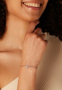 Verstelbare zilveren armband met een roze bloemencharme, met een delicate ketting en een gepolijste afwerking tegen een neutrale stoffen achtergrond.