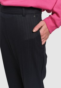 Czarne spodnie w paski wykonane z gładkiego materiału, z kieszenią boczną i małym metalowym logo przypiętym do pasa.