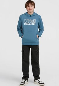 Modra kapa z belim napisom logotipa, sprednjim žepom in kapuco z vrvico. Črne hlače cargo z bočnimi žepi. Bele superge s črnimi poudarki.