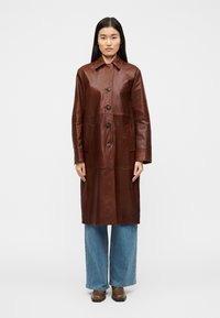 Καφέ δερμάτινο παλτό με γιακά, εμπρόσθιο κούμπωμα με κουμπιά, δύο πλαϊνές τσέπες και μήκος μέχρι τη μέση. Φοριέται πάνω από φαρδιά μπλε τζιν.