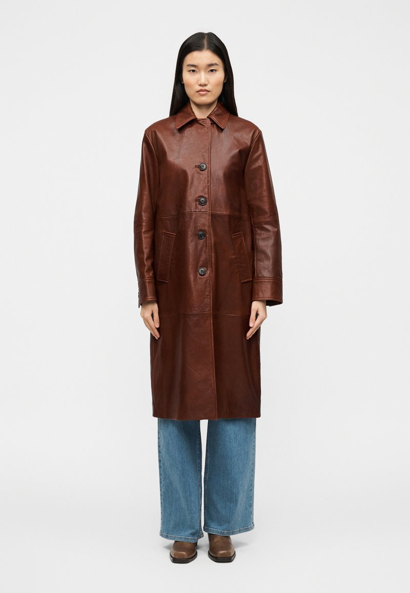 Καφέ δερμάτινο παλτό με γιακά, εμπρόσθιο κούμπωμα με κουμπιά, δύο πλαϊνές τσέπες και μήκος μέχρι τη μέση. Φοριέται πάνω από φαρδιά μπλε τζιν.