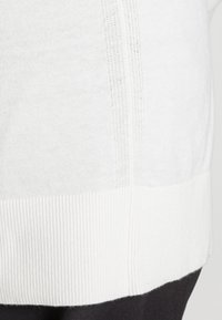 Vit tröja med ribbad nederkant och texturerade sidopaneler. Slät tyg med subtilt mönster. Minimalistisk design, mjuk textur.