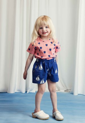 Il bambino indossa una maglietta rosa a costine con motivi a cuore blu e pantaloni corti blu con stampe di velieri bianche, abbinati a zoccoli bianchi.