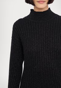 Femme portant un pull à col roulé côtelé noir avec de subtils fils métalliques et une coupe au carré droite, montrée des épaules aux lèvres.