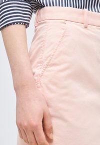 Približen posnetek osebe, ki nosi bledo rožnate hlače, s roko, ki počiva blizu sprednjega žepa, in dolgimi rokavi modro-bele črtaste majice.