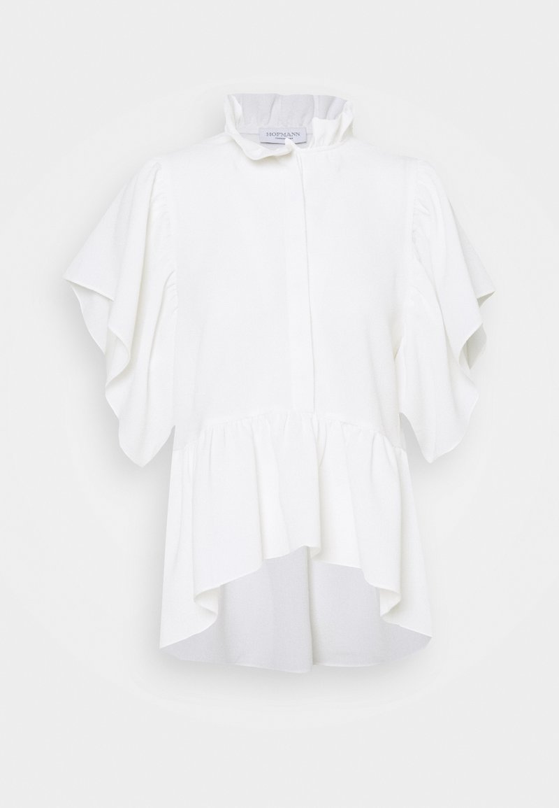 Blouse blanche avec col montant volanté, manches papillon et ourlet basque asymétrique, présentée sur un fond clair uni.