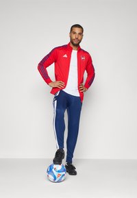 Rød atletisk jakke med blå striber, hvid T-shirt, marineblå træningsbukser med hvide sidelister og sorte sneakers; stående på en fodbold.