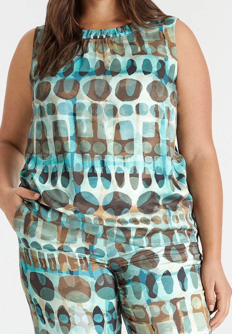 Top sans manches et pantalon avec un motif géométrique abstrait bleu, marron et teal porté par une personne avec la main dans la poche.