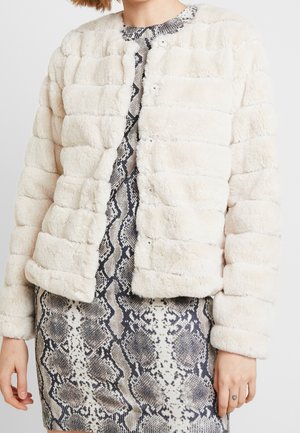 Femme portant une veste matelassée blanche et duveteuse par-dessus une robe noire et blanche à motif peau de serpent, avec un petit tatouage en forme de cœur sur son doigt.