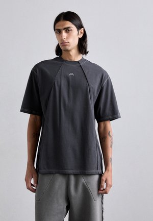 Camiseta de manga corta en gris oscuro, hecha de algodón suave. Presenta mangas raglán y detalles de costura, con un pequeño logo en la parte delantera.