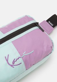 Borsa multicolore con tessuto verde chiaro e viola, dotata di una cerniera nera e un logo ricamato in filo viola. Texture morbida.