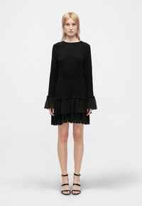 Vestido negro con mangas largas y un dobladillo en capas y acampanado. Tejido texturizado con acentos de volantes. Combinado con sandalias negras con tiras.