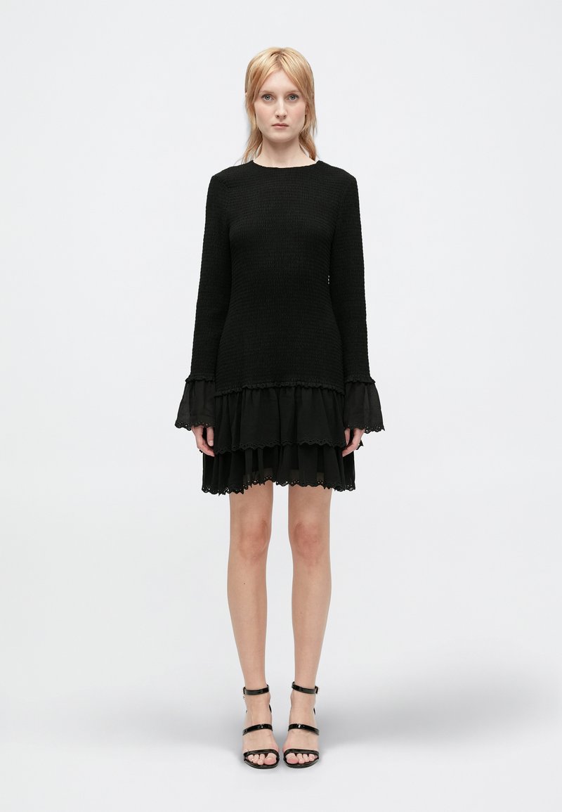 Vestido negro con mangas largas y un dobladillo en capas y acampanado. Tejido texturizado con acentos de volantes. Combinado con sandalias negras con tiras.