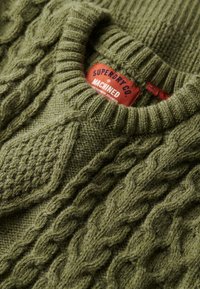 Zielony sweter z dzianiny z wzorem w warkocz, ze żebrowanym kołnierzem i metką Superdry Co. Teksturowana powierzchnia z różnorodnymi wzorami dzianiny.