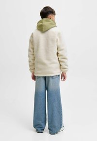 Witte schapenvachtjas met een groene capuchon, gecombineerd met lichtblauwe jeans met wijde pijpen. De outfit heeft casual, relaxte vormen en texturen.