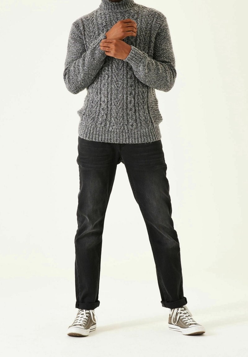 Pull à col roulé gris en maille avec un motif texturé, associé à un jean en denim noir. Des baskets basses grises et blanches complètent la tenue.