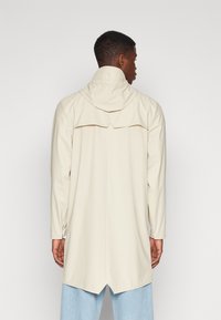 Beige regnfrakk med hette, lange ermer og bakdetalj, laget av glatt, vanntett materiale, med ventilert nederkant.