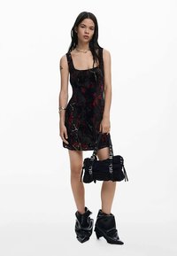 Robe à motifs noirs et rouges avec un décolleté carré, associée à un sac noir à texture plissée et ferrures sur la bandoulière, portée avec des bottes noires brillantes.