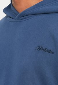 Blå hettegenser laget av mykt stoff har en stor hette og en subtil brodert logo på brystet i en mørkere blånyanse.