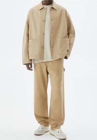 Beige bomuldsjakke med krave og to forlommer, kombineret med en lysebeige t-shirt og matchende bukser med vidde i benene. Hvide sneakers.