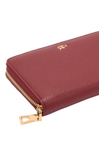 Portfel z bordowej skóry o fakturowanym wykończeniu, z zamkiem błyskawicznym w złotym odcieniu i małym logo wytłoczonym z przodu. Suwak zamka ma nazwę marki.