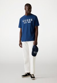Tummansininen puuvillapaita, jossa on valkoiset "GUESS EST. 1981" -tekstit, yhdistetty valkoisiin housuihin ja tummansiniseen baseball-lippikseen. Mustat lenkkarit vaaleanbeigeillä yksityiskohdilla.