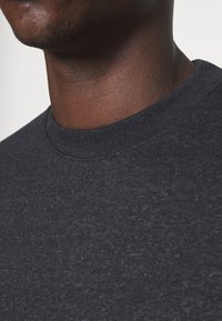 Närbild av en persons nacke och övre bröst som bär en mörk heather-färgad Nike sweatshirt med en diskret logotyp på vänster sida.