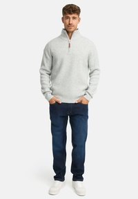 Mand står med hænderne i lommerne iført en lysegrå zip-sweater, mørkeblå jeans og hvide sneakers på en hvid baggrund.