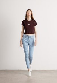 Czarny krótki t-shirt z logo, jasnoniebieskie obcisłe dżinsy i białe sneakersy. Model stoi na tle jednolitym, prezentując strój.