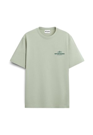 Osoba z kręconymi blond włosami nosi jasnzieloną koszulkę z rysunkiem filiżanki herbaty i sztućców oraz napisem „Matcha Friends Team Green Supreme.”