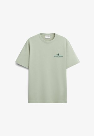 Osoba z kręconymi blond włosami nosi jasnzieloną koszulkę z rysunkiem filiżanki herbaty i sztućców oraz napisem „Matcha Friends Team Green Supreme.”
