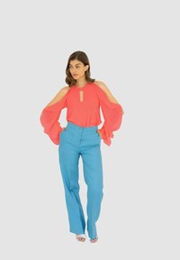 Blusa corallo con design a spalla scoperta e dettaglio a goccia, abbinata a pantaloni a gamba larga a vita alta di colore azzurro chiaro. Tessuti lisci, senza motivi.