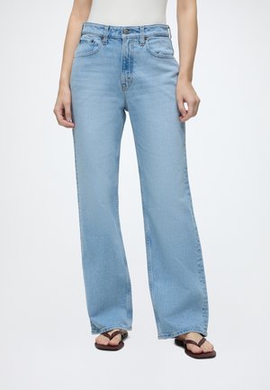 Personne portant un jean droit taille haute bleu clair et des sandales tongs marron foncé, debout devant un fond blanc.