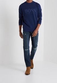 Mörkblå crewneck sweatshirt med "BOSS"-logotyp, kombinerad med slitna blå jeans och bruna mockastövlar, med en avslappnad passform.