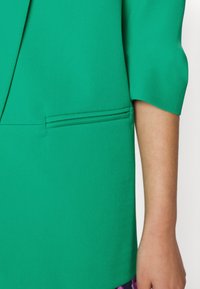 Zelený blejzr s hladkým materiálem, štíhlým střihem a jemným detailem kapsy na levé přední straně. Krátké rukávy.