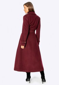 Abrigo largo de lana burdeos con cintura ceñida, cuello amplio y puños abotonados. Textura suave y diseño hasta el suelo.