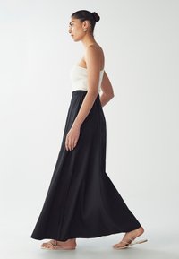 Gonna maxi nera con una texture liscia, abbinata a un top bianco senza maniche a coste. Il modello indossa sandali con cinturini sottili.