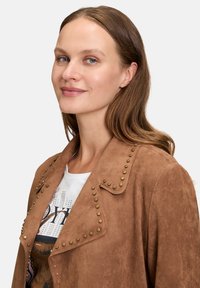 Veste en daim marron avec des accents cloutés le long du col et sur le devant, associée à un t-shirt graphique blanc. Texture lisse et design structuré.