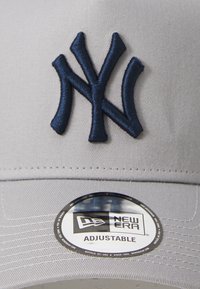 Grå baseballcap med marineblå brodert New York Yankees-logo. Har justerbar stropp og sirkulært New Era-merke på skyggen.