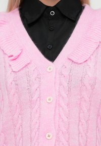 Ανοιχτό ροζ πλεκτό ζακέτα με σχέδια από καλώδιο, V-λαιμό και ριχτό γιακά. Διαθέτει λευκά κουμπιά και στρώσεις με ρίγες στην περιοχή του λαιμού.