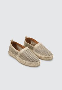 Par de alpargatas beige sin cordones con parte superior perforada y suela trenzada de yute sobre un fondo claro y liso.