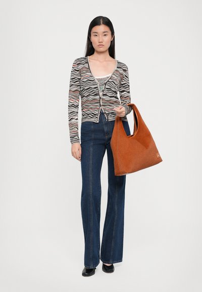 Cardigan à motifs avec un design en vagues noir, blanc et rose, associé à un jean bleu évasé, tenant un sac à main en feutre brun.