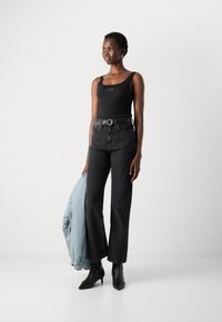 Débardeur noir avec logo, jean taille haute noir, bottes à talons noires et une veste bleu clair portée sur le bras.