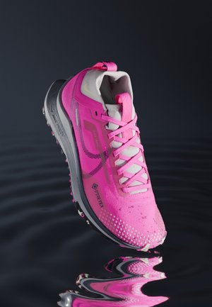 Jasnoróżowy but do biegania z białymi sznurowadłami, oznaczeniem Gore-Tex i szarą podeszwą unoszący się nad odbijającą ciemną wodą.