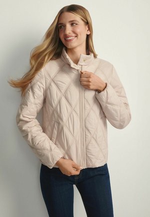 Jasnoróżowa pikowana kurtka z wysokim kołnierzem, ukośnym wzorem szwów, z przodu zamknięcie na zamek błyskawiczny i długie rękawy. Do tego ciemne dżinsy.