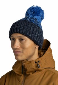 Knit beanie with a blue and navy gradient, featuring a large blue pom-pom on top and ribbed texture. Worn with a brown jacket.