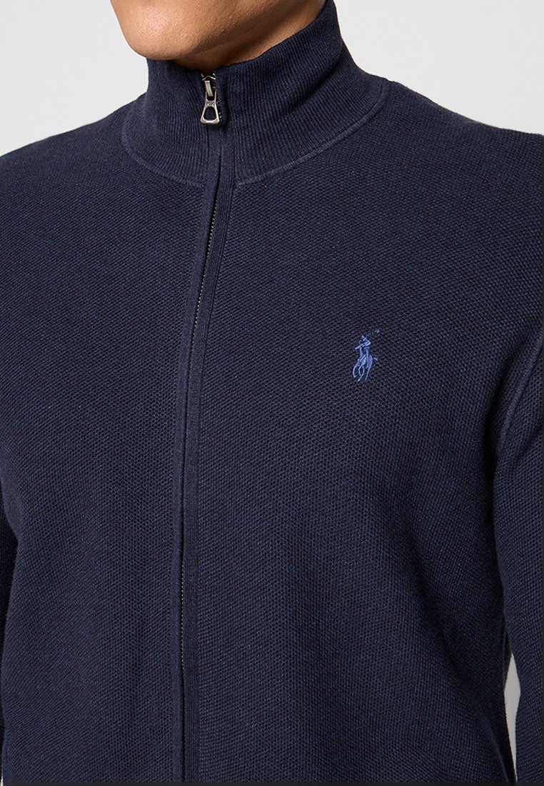 Marinefarbener Zip-Pullover mit hohem Kragen, strukturiertem Strickstoff und kleinem blauen bestickten Logo auf der linken Seite. Der Reißverschluss ist sichtbar.