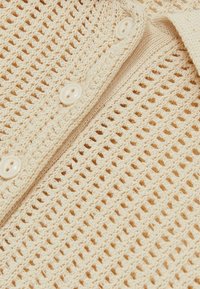 Top lavorato a maglia beige con un design a rete, con piccoli fori quadrati e bottoni in plastica bianchi lungo il patta. Tessuto morbido e leggero.