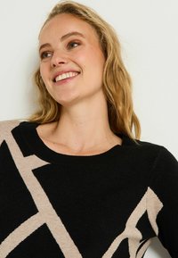 Pull à motifs noirs et beiges avec un col rond, présentant des motifs angulaires aux couleurs contrastées et un tricot doux et texturé.